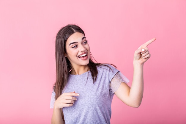 Portret van een brunette vrouw met lang haar in een basisch t-shirt die zich verheugt en met de vinger naar copyspace wijst, geïsoleerd op een roze achtergrond