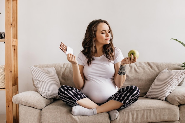 Portret van een brunette zwangere vrouw in een wit T-shirt die probeert te kiezen tussen een gezonde, groene appel en smakelijke melkchocolade Meisje zit op een beige bank