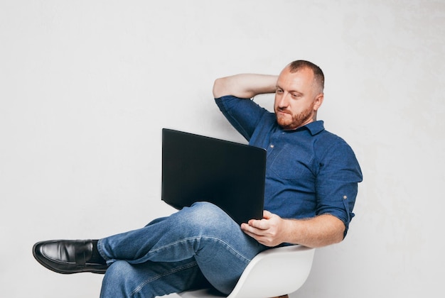 Portret van een brutale man zittend op een stoel tegen een witte muur met een laptop en een kladblok zakenman