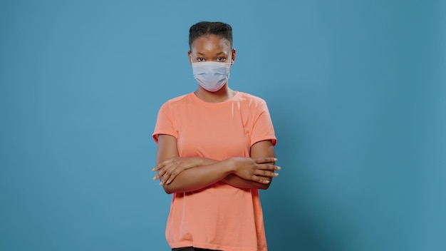 Portret van een casual vrouw die een beschermend masker op het gezicht zet terwijl ze naar de camera kijkt. Jongere die bescherming heeft tegen coronavirus en staat met armen gekruist over blauwe achtergrond.