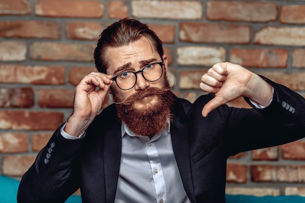 Portret van een charmante aantrekkelijke man met bril, snor en baard in een pak, kijkend naar de camera en met zijn duimen naar beneden.