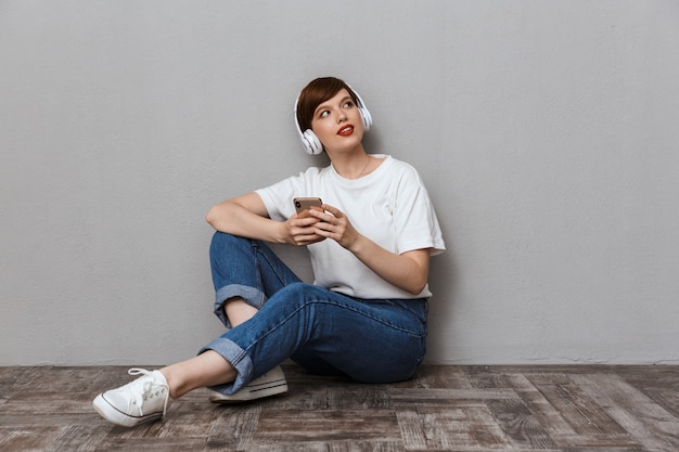 Portret van een charmante jonge vrouw die mobiele telefoon en koptelefoon gebruikt terwijl ze op de vloer zit geïsoleerd over een grijze muur