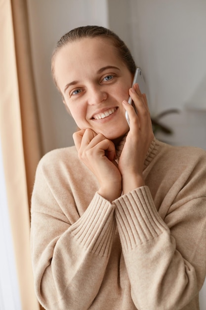 Portret van een charmante, schattige jonge volwassen vrouw met een beige trui die op een mobiele telefoon praat en de hand op de kin houdt en naar de camera kijkt met een vriendelijke brede glimlach