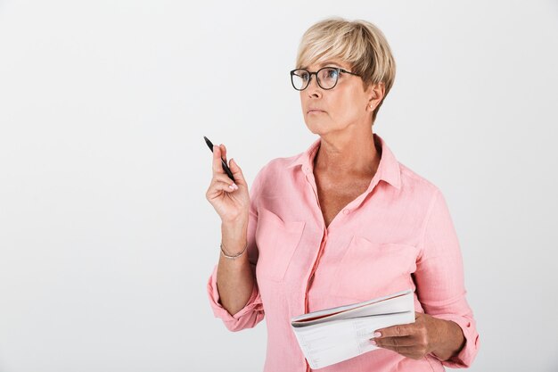 Portret van een charmante volwassen vrouw met een bril met een studerend boek en een pen geïsoleerd over een witte muur in de studio