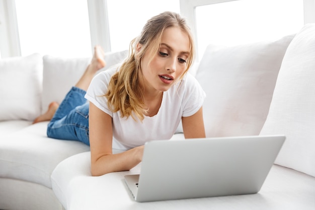 Portret van een charmante vrouw van 20 jaar die een zilveren laptop gebruikt terwijl ze thuis op de bank in de woonkamer ligt