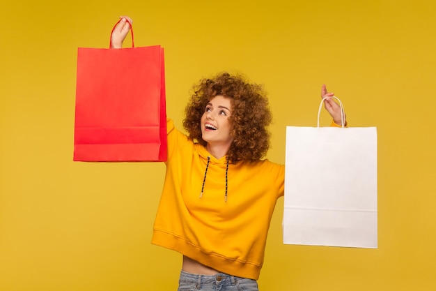 Portret van een dolblij gelukkig hipstermeisje met krullend haar in hoodie die boodschappentassen opheft met een leeg gebied, kopieer ruimte op pakketten voor reclametekst. indoor studio-opname, commercial van modewinkel