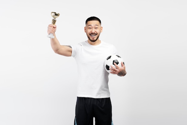 Portret van een dolblije aziatische voetballer die een bal vasthoudt en een trofeebeker viert