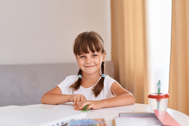 Portret van een donkerharig meisje dat iets tekent in een album dat aan de tafel bij het raam zit, schattig kind met vlechten die naar de camera kijkt met een vrolijke gezichtsuitdrukking