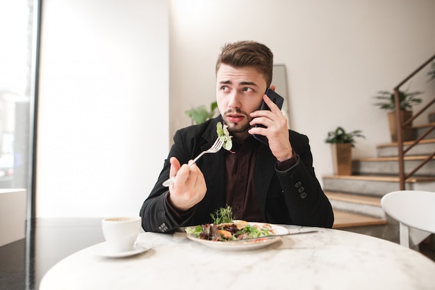 Portret van een drukke man die praat aan de telefoon en eet een salade met een vork in een gezellig restaurant, wegkijken