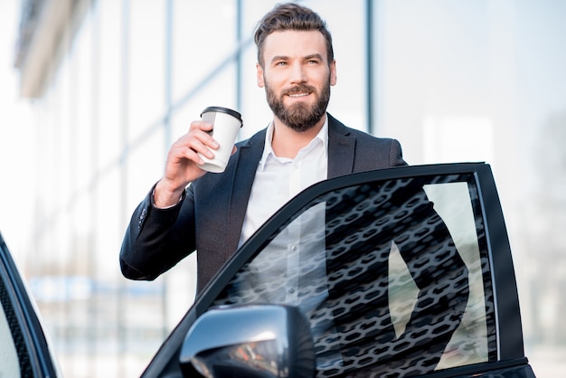 Portret van een elegante zakenman die met een koffiekopje in de buurt van de auto staat