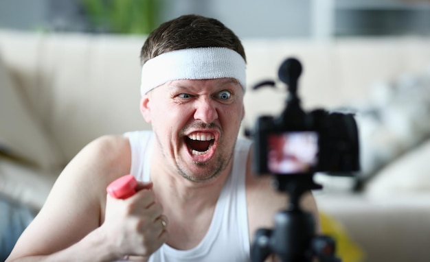Portret van een emotioneel mannelijke blogger met een grimace op het gezicht en met dumbbells voor de hand