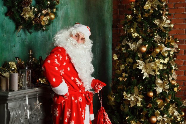 Portret van een emotionele kerstman met een zak geschenken bij de kersthaard in een ingerichte woonkamer met kerstboom. De kerstman komt 's avonds en brengt cadeautjes. Ruimte voor site kopiëren