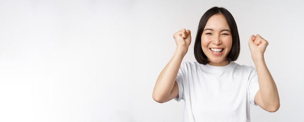 Portret van een enthousiaste aziatische vrouw die vieren en triomferen wint en de handen omhoog steekt om een doel of succes te bereiken dat op een witte achtergrond staat