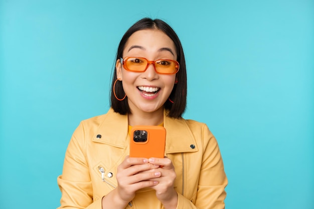 Portret van een enthousiaste aziatische vrouw met een zonnebril die een mobiele telefoon gebruikt die lacht en lacht en er gelukkig uitziet met een smartphone die over een blauwe achtergrond staat