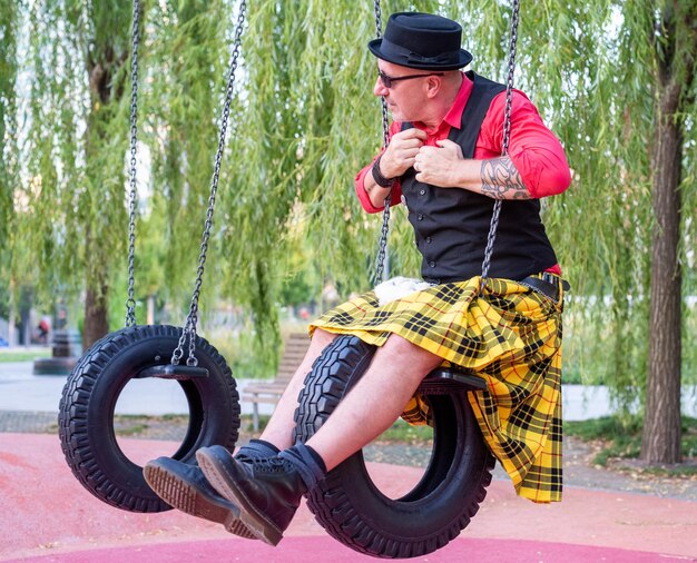 Foto portret van een excentrieke man gekleed in een kilt spelen op schommels gemaakt van banden volwassene in kinderhouding in een speeltuin onvolwassenheid concept