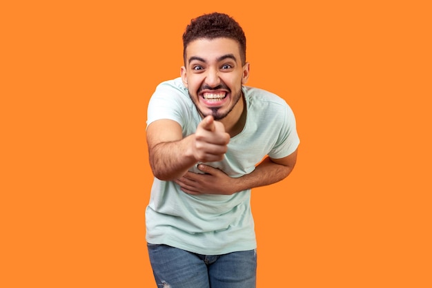 Portret van een extreem gelukkige brunette man met baard in wit t-shirt hand op buik, wijzend op de camera en hardop lachend, je belachelijk makend. studio-opname geïsoleerd op oranje achtergrond