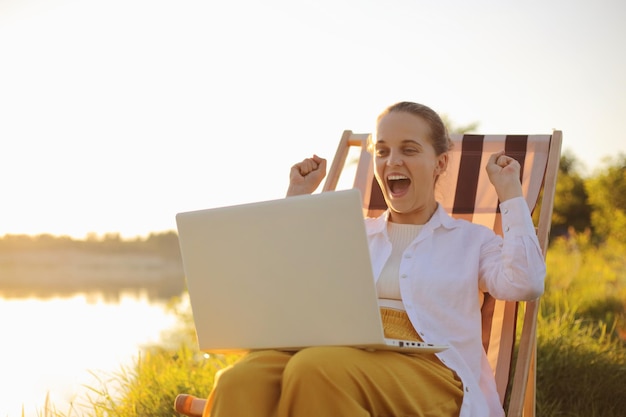 Portret van een extreem gelukkige positieve vrouw met een wit overhemd aan het water zittend in een klapstoel en bezig met laptop wedden en winnen tijdens vakantie met opgeheven armen en schreeuwend