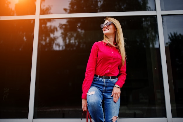 Portret van een fantastische jonge vrouw in rode blouse en spijkerbroek poseren met haar handtas en zonnebril buiten het winkelcentrum