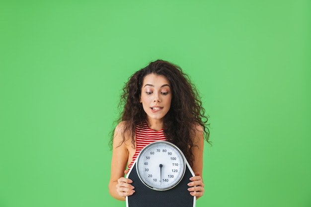 Portret van een fitnessvrouw uit de twintig die zomerkleren draagt met een weegschaal tijdens oefeningen op de groene muur