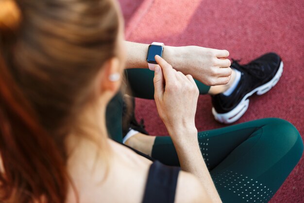 Foto portret van een fitte jonge vrouw die een trainingspak draagt en naar haar polshorloge kijkt terwijl ze op het sportveld zit tijdens de training in het groene park