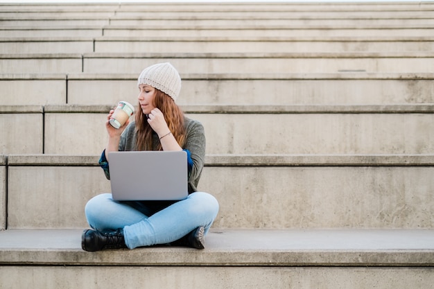 Portret van een freelancer-vrouw die een laptop gebruikt en koffie drinkt terwijl ze op de trap in de stadsstraat zit
