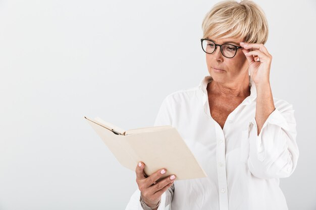 Portret van een geconcentreerde volwassen vrouw met een bril die een boek leest dat over een witte muur in de studio wordt geïsoleerd