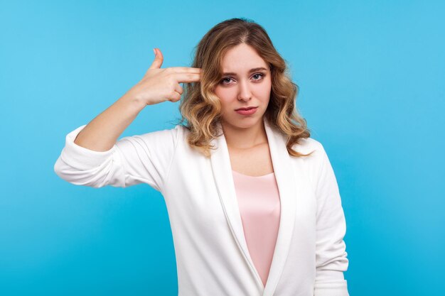 Portret van een gefrustreerde vrouw met golvend haar in een witte jas wijzend met een vingerpistool naar het hoofd met een verveelde gezichtsuitdrukking, zichzelf neerschietend in depressie, moe van het leven. indoor studio-opname, blauwe achtergrond