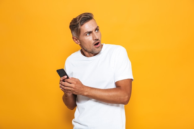 Portret van een geïrriteerde man van 30 in een wit t-shirt die een mobiele telefoon vasthoudt en praat terwijl hij geïsoleerd op geel staat