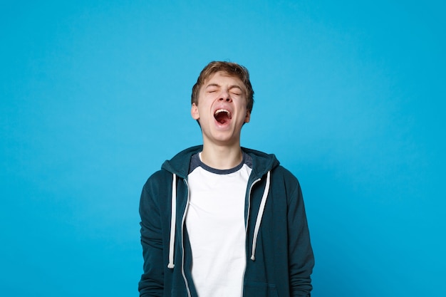 Portret van een gekke jongeman in vrijetijdskleding die de ogen gesloten houdt en schreeuwend geïsoleerd op een blauwe muur.