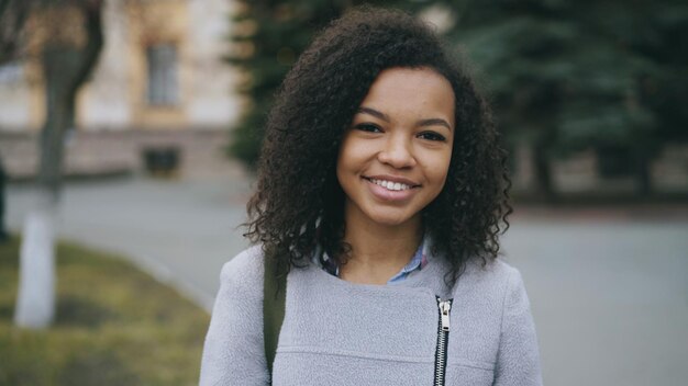 Portret van een gekruld studentenmeisje van gemengd ras dat in de camera glimlacht en lacht om de straat van de stad