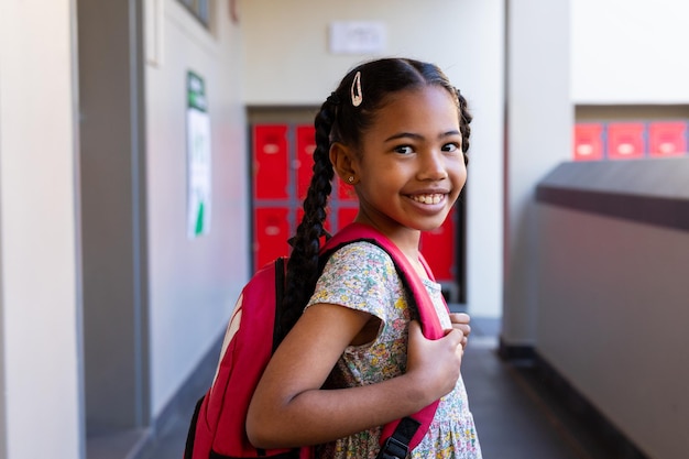 Portret van een gelukkig biracial schoolmeisje met een glimlachende schooltas in de gang van de basisschool