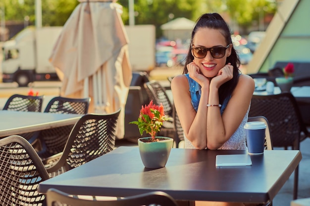 Portret van een gelukkig charmant brunette meisje dat trendy kleding draagt, geniet van een zomerdag terwijl ze op een terras in het openluchtcafé zit en naar de camera kijkt.