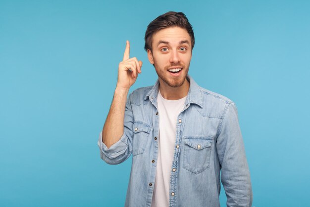 Portret van een gelukkig geïnspireerde man in een werknemer-denim-shirt die de vinger opsteekt en een geniaal idee heeft, verbaasd door een plotselinge slimme oplossing, creatieve gedachte. indoor studio-opname geïsoleerd op blauwe achtergrond