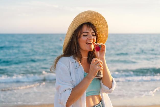 Portret van een gelukkig jong meisje in een strohoed met een cocktail in de hand op een achtergrond van zee