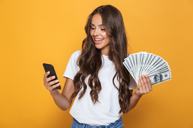 Portret van een gelukkig jong meisje met lang donkerbruin haar dat zich over gele muur bevindt, die geldbankbiljetten houdt, die mobiele telefoon gebruikt