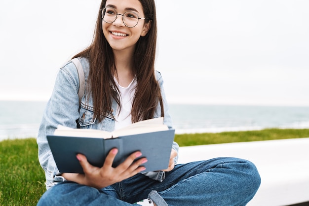 Portret van een gelukkig jong tienermeisje met een bril die lacht en een boek leest terwijl ze op groen gras aan zee zit