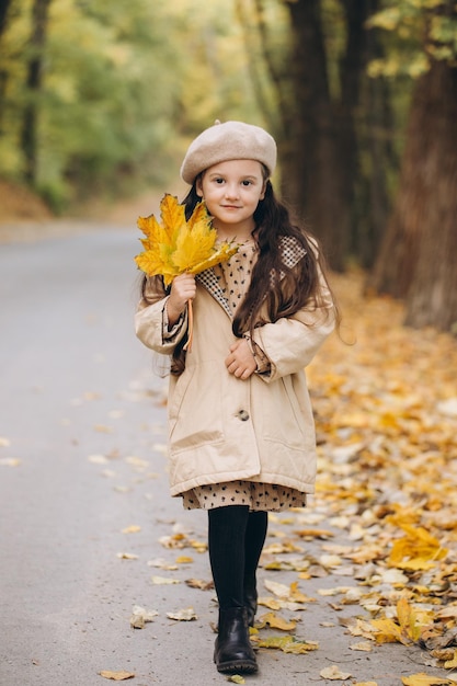 Portret van een gelukkig klein meisje in een beige jas en baret die gele esdoornbladeren vasthoudt en tijd doorbrengt in het herfstpark