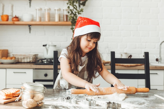 Portret van een gelukkig klein meisje in een kerstmuts die deeg op de keukentafel uitrolt, een kind dat kerstkoekjes bereidt