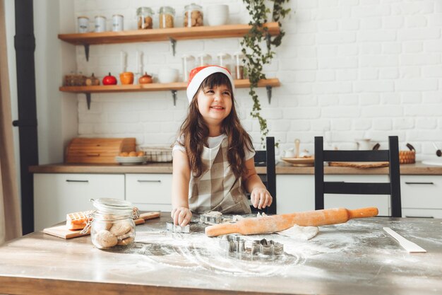 Portret van een gelukkig klein meisje in een kerstmuts die deeg op de keukentafel uitrolt, een kind dat kerstkoekjes bereidt