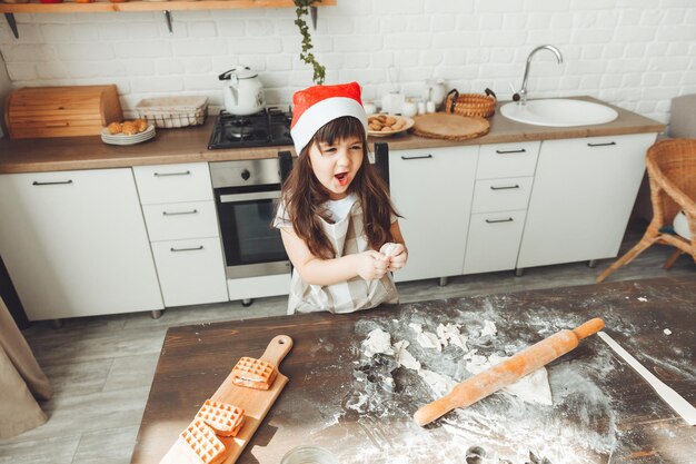 Portret van een gelukkig klein meisje in een kerstmuts die deeg op de keukentafel uitrolt, een kind dat kerstkoekjes bereidt