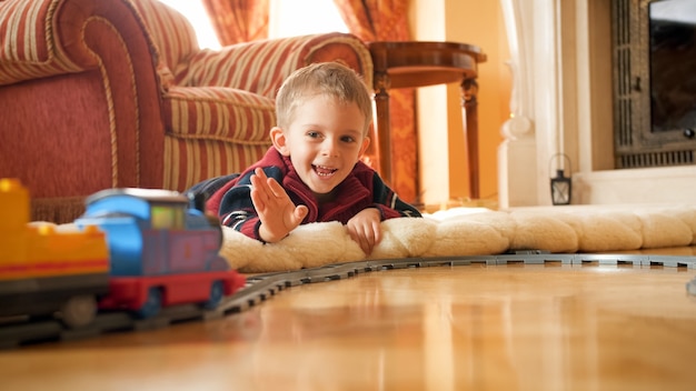 Portret van een gelukkig lachend jongetje dat op de vloer ligt en met de hand zwaait naar speelgoedtrein die op de spoorlijn in de woonkamer rijdt.