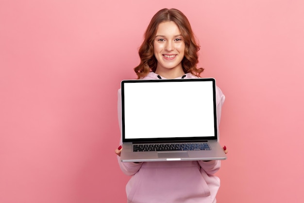 Portret van een gelukkig lachend tienermeisje met krullend haar in hoodie met een leeg laptopscherm op de camera, plaats voor reclame. Indoor studio-opname geïsoleerd op roze background