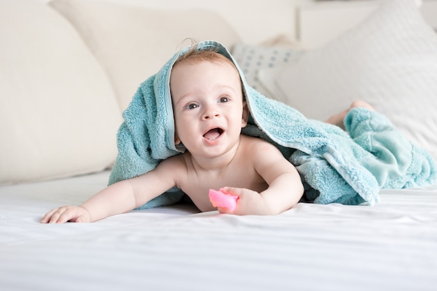 Portret van een gelukkig lachende baby onder een blauwe handdoek die op bed kruipt met witte lakens