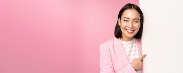 Portret van een gelukkig lachende Japanse zakelijke vrouw die met de vinger naar het bord wijst en iets laat zien op lege ruimte in pak over roze achtergrond