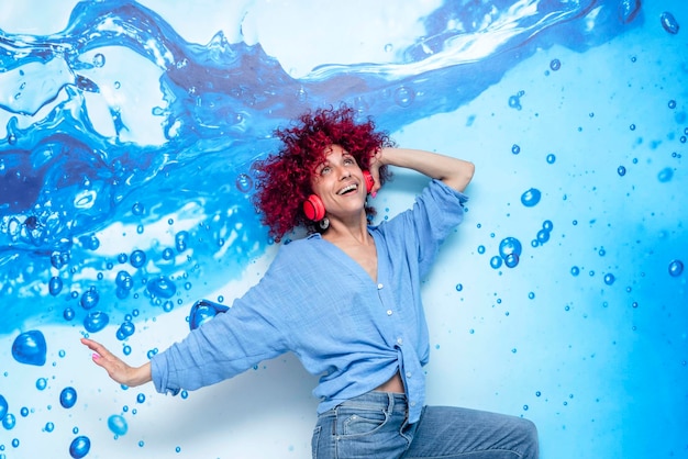 Portret van een gelukkig lachende jonge latijnse vrouw met rood afro haar die plezier heeft met dansen en luisteren naar muziek en op haar rode koptelefoon op een blauwe achtergrond met blauwe druppels