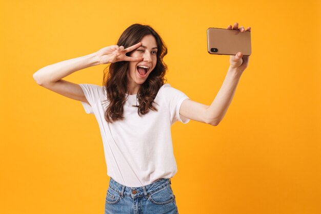 Portret van een gelukkig lachende jonge vrouw die zich geïsoleerd over een gele muur bevindt, een selfie maakt met de mobiele telefoon en een vredesgebaar maakt