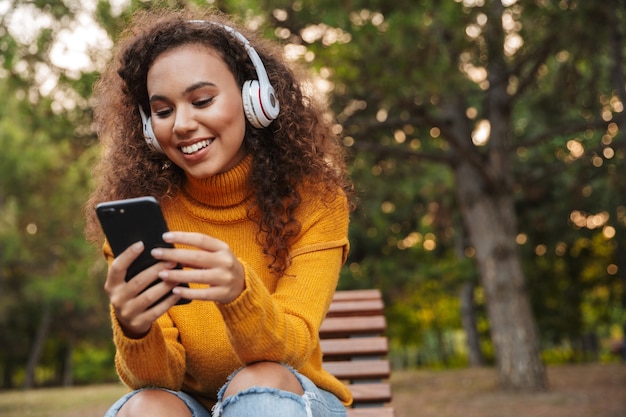 Portret van een gelukkig lachende mooie jonge krullende vrouw zit op een bankje in het park buiten en luistert naar muziek met een koptelefoon via de mobiele telefoon.