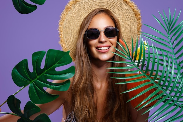 Portret van een gelukkig meisje met een strohoed in bikini en een zonnebril die tussen tropische planten staat