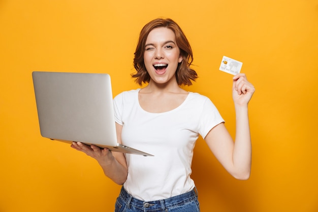 Portret van een gelukkig mooi meisje dat geïsoleerd over een gele muur staat, met behulp van een laptopcomputer, met creditcard
