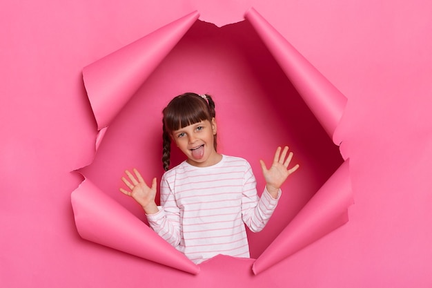 Portret van een gelukkig positief grappig klein meisje met een gestreept shirt poseren in roze papier gescheurd staande met opgeheven armen en tong uit met plezier fronsend gezicht